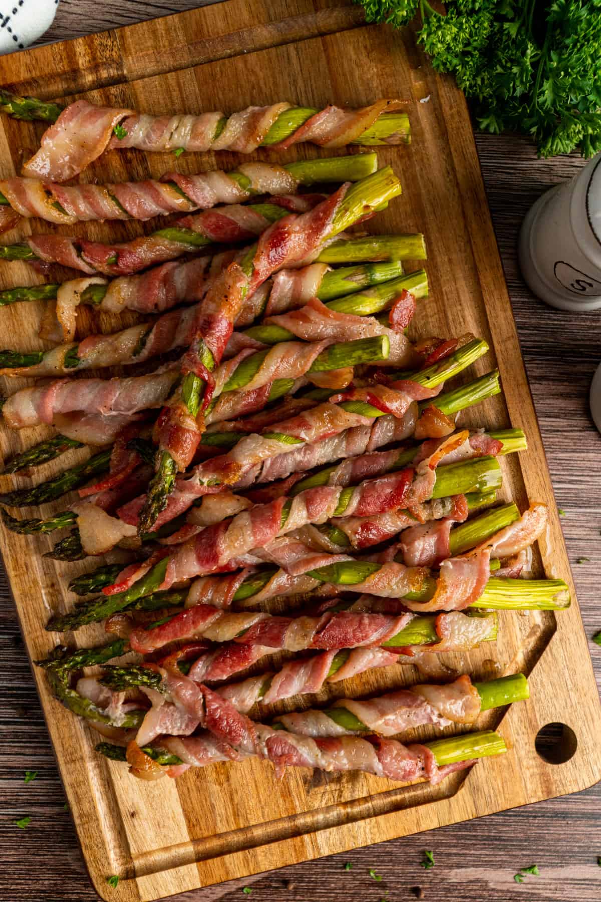 Asparagus wrapped in bacon piled up on a wood cutting board.