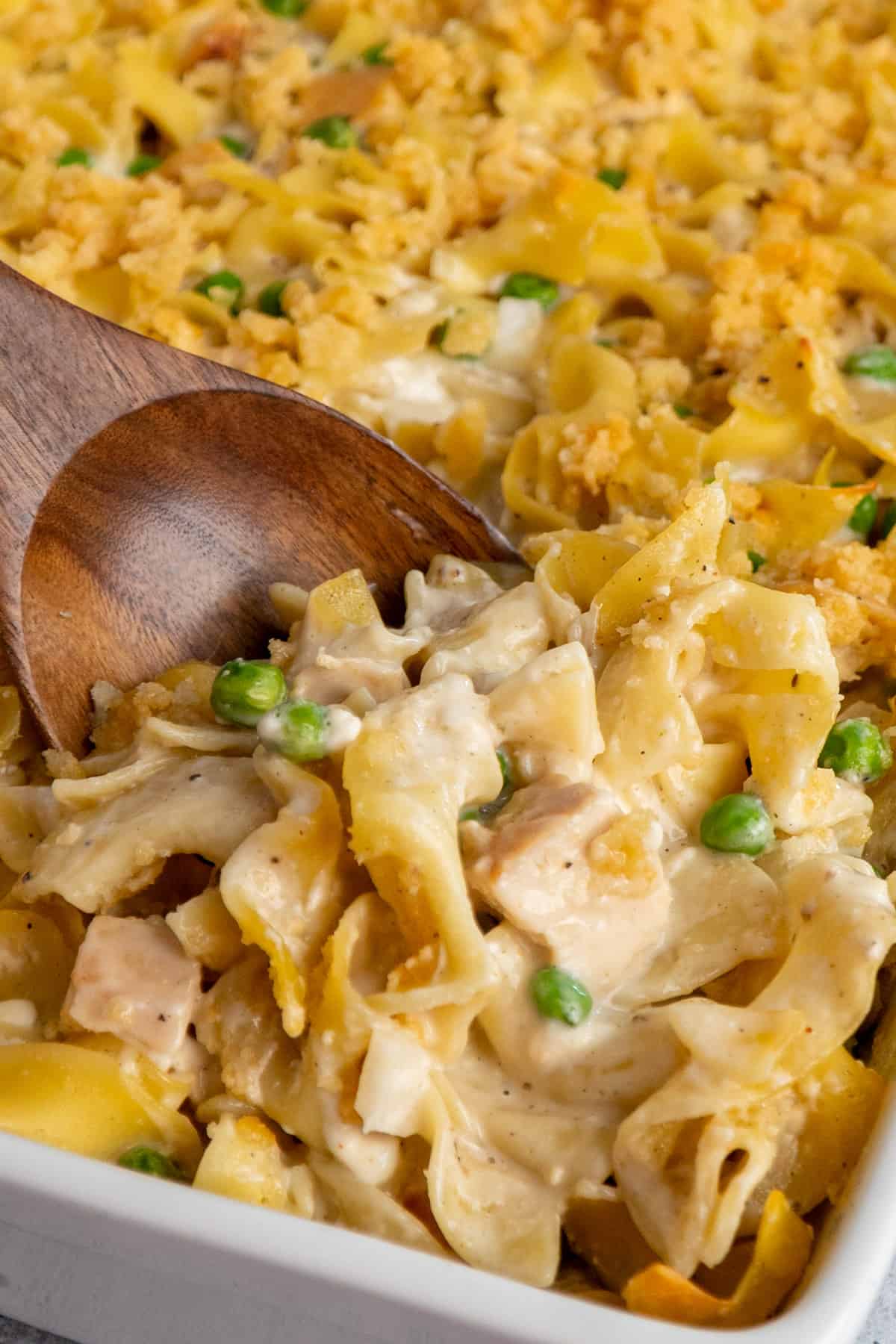Serving chicken noodle casserole from a white baking dish with a wooden spoon.