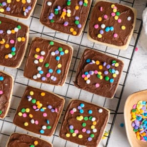 Chocolate frosted shortbread cookies on a cooling rack with sprinkles.