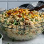 Close-up of pea pasta salad in a clear bowl with a wooden spoon in it.