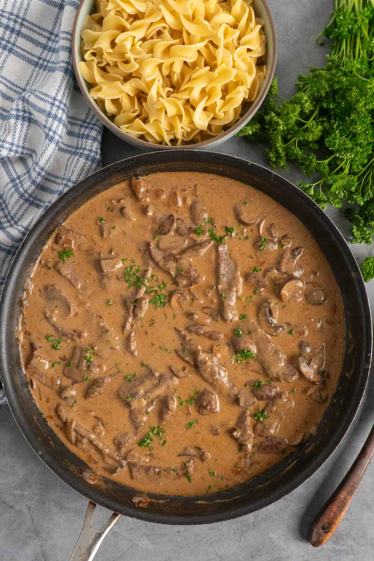 Beef stroganoff in a skillet next to a bunch of parsley and bowl of egg noodles.