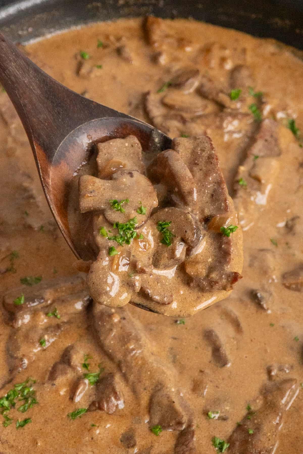 A wooden spoon holding a scoop of beef stroganoff up over the pan of more.