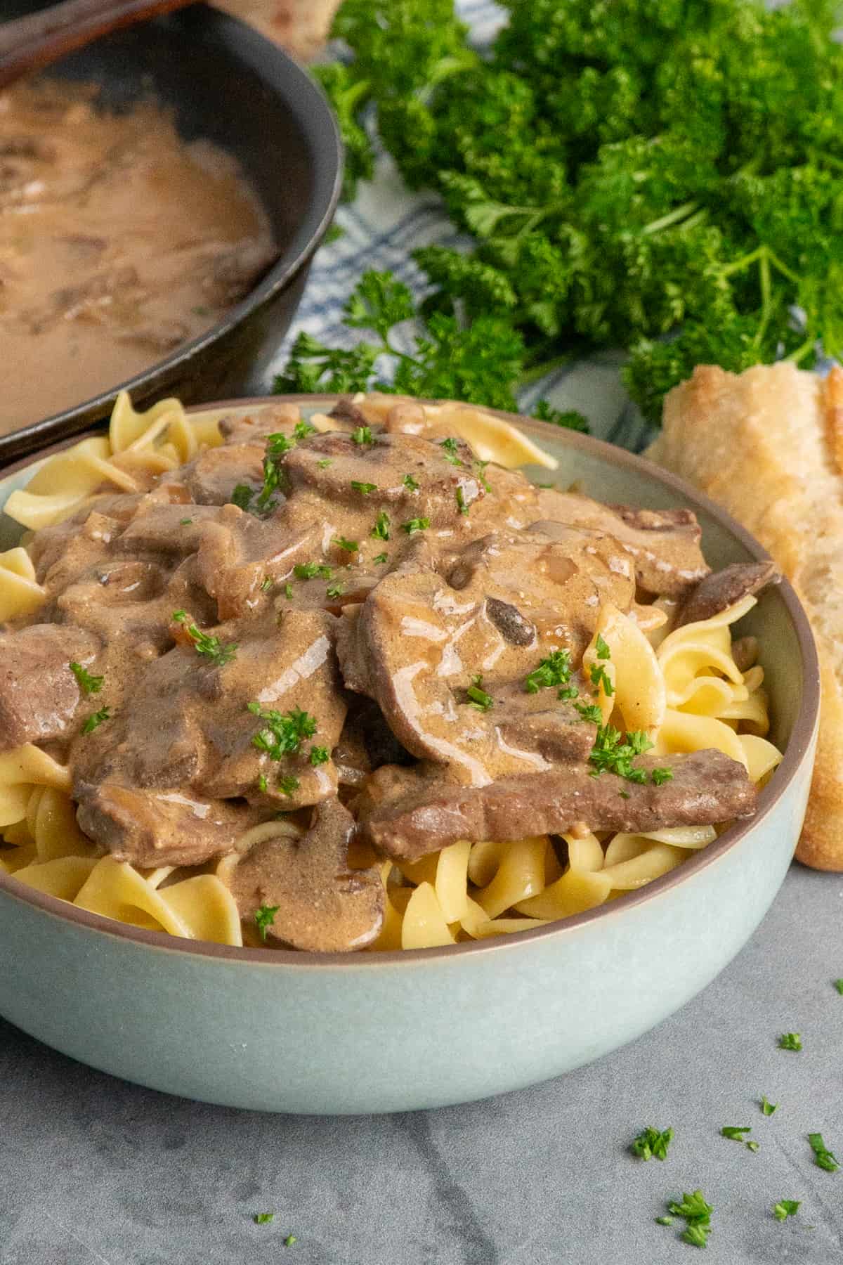 Beef stroganoff over a bowl of egg noodles with parsley and some bread in the background.