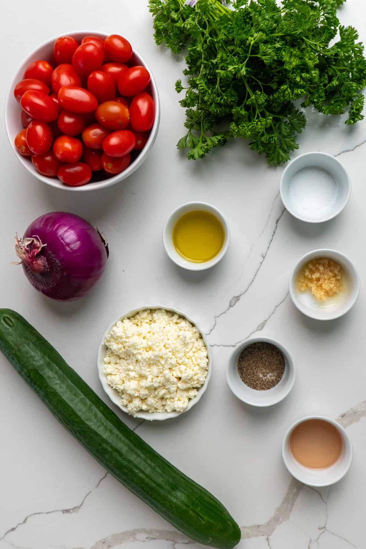 All the ingredients to make cucumber tomato salad on a white marble counter in bowls.