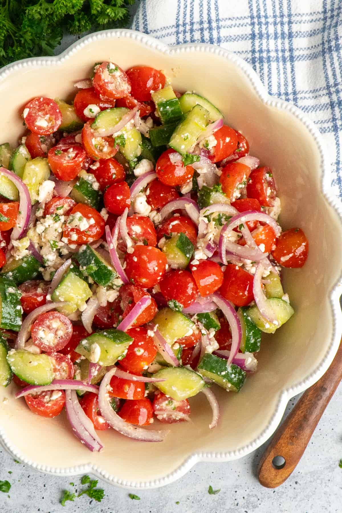 A bowl of cucumber and tomato salad on a table with a blue and white tea towel and parsley on the side.