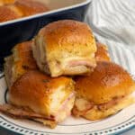 Five ham and cheese sliders on a plate with a blue baking dish in the background.