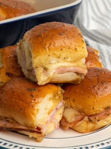 Five ham and cheese sliders on a plate with a blue baking dish in the background.