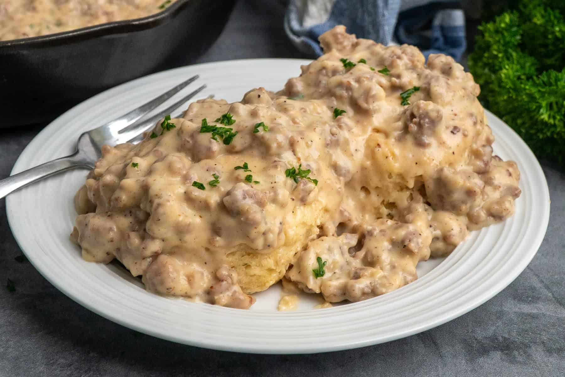 Biscuits on a white plate covered in sausage and gravy with a skillet in the background.