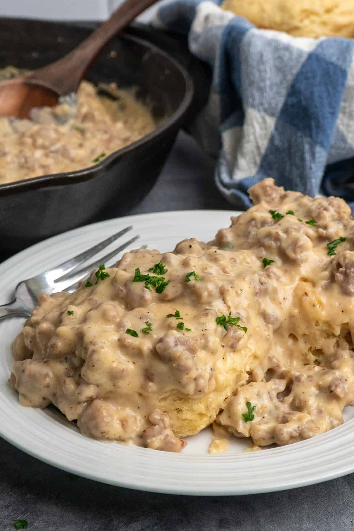 Biscuits on a white plate covered in sausage and gravy with a skillet in the background.