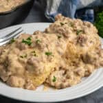 Biscuits on a white plate covered in sausage and gravy with a skillet in the background.