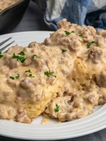 Biscuits on a white plate covered in sausage and gravy with a skillet in the background.