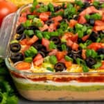 A glass dish of 7 layer taco dip with the different layers visible and colorful toppings on the top.