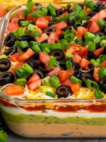 A glass dish of 7 layer taco dip with the different layers visible and colorful toppings on the top.