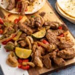 Air fryer steak fajitas and vegetables resting on wooden board with tortillas to the side.