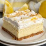 A lemon cheesecake bar on a plate with lemons in the background.