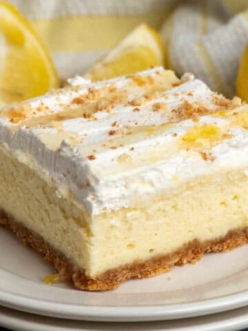 A lemon cheesecake bar on a plate with lemons in the background.