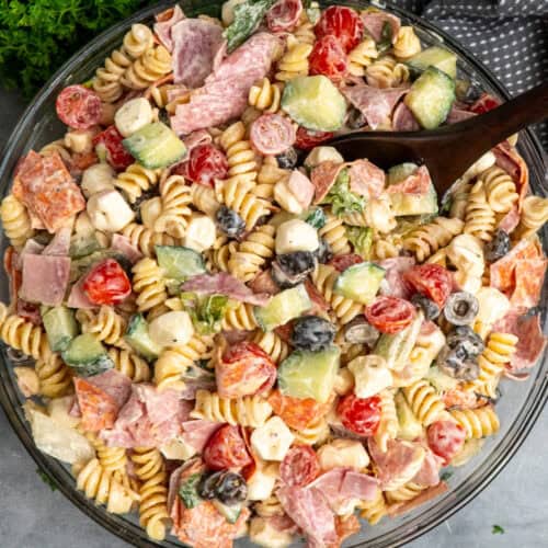 Italian grinder pasta salad in a clear bowl with a wooden spoon in it.