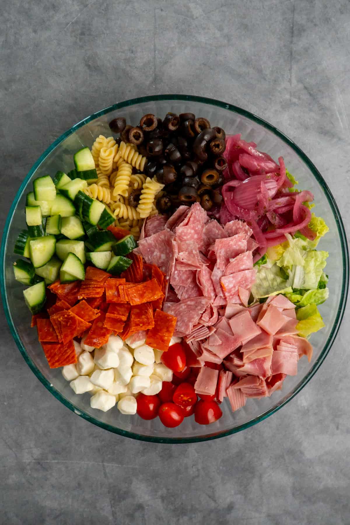 Pasta, slice meats mozzarella pearls, and veggies in a clear bowl.