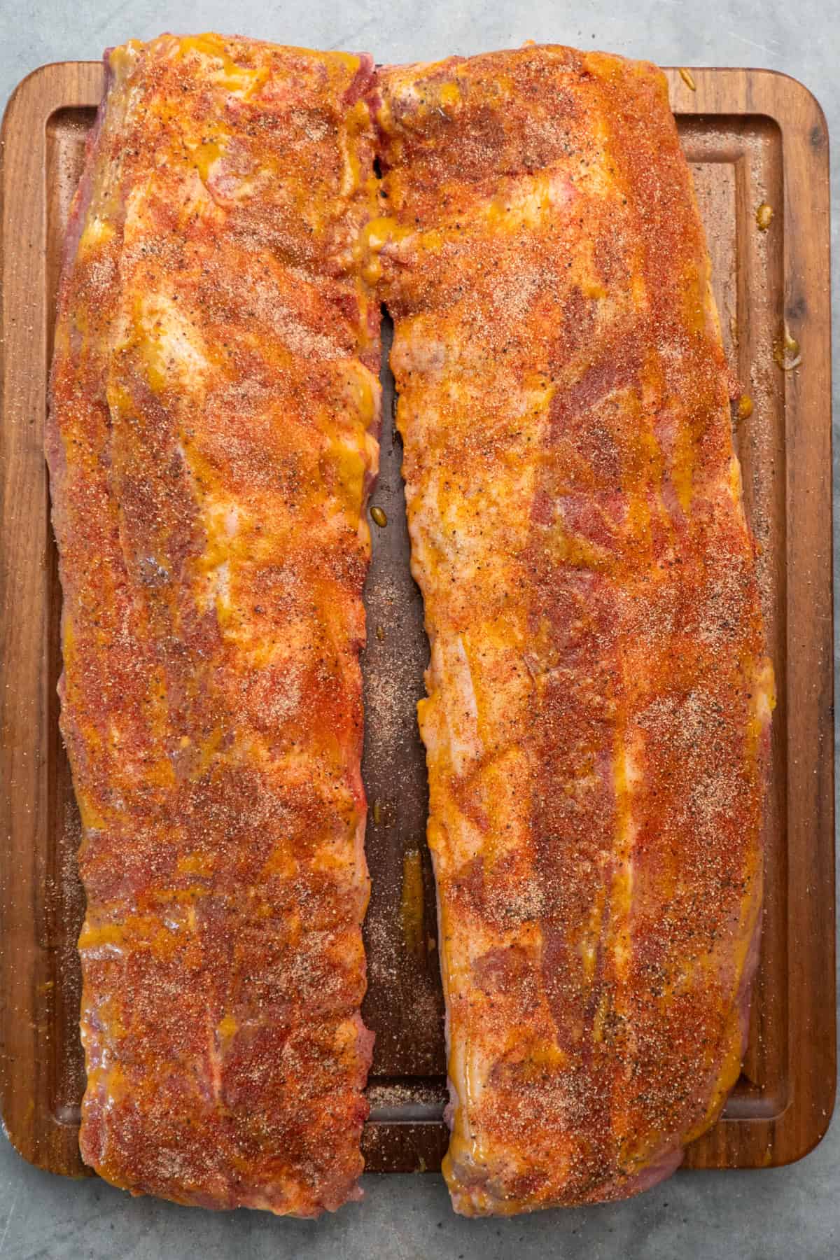 Pork ribs covered in mustard sauce and dry rub seasonings on a wood cutting board.
