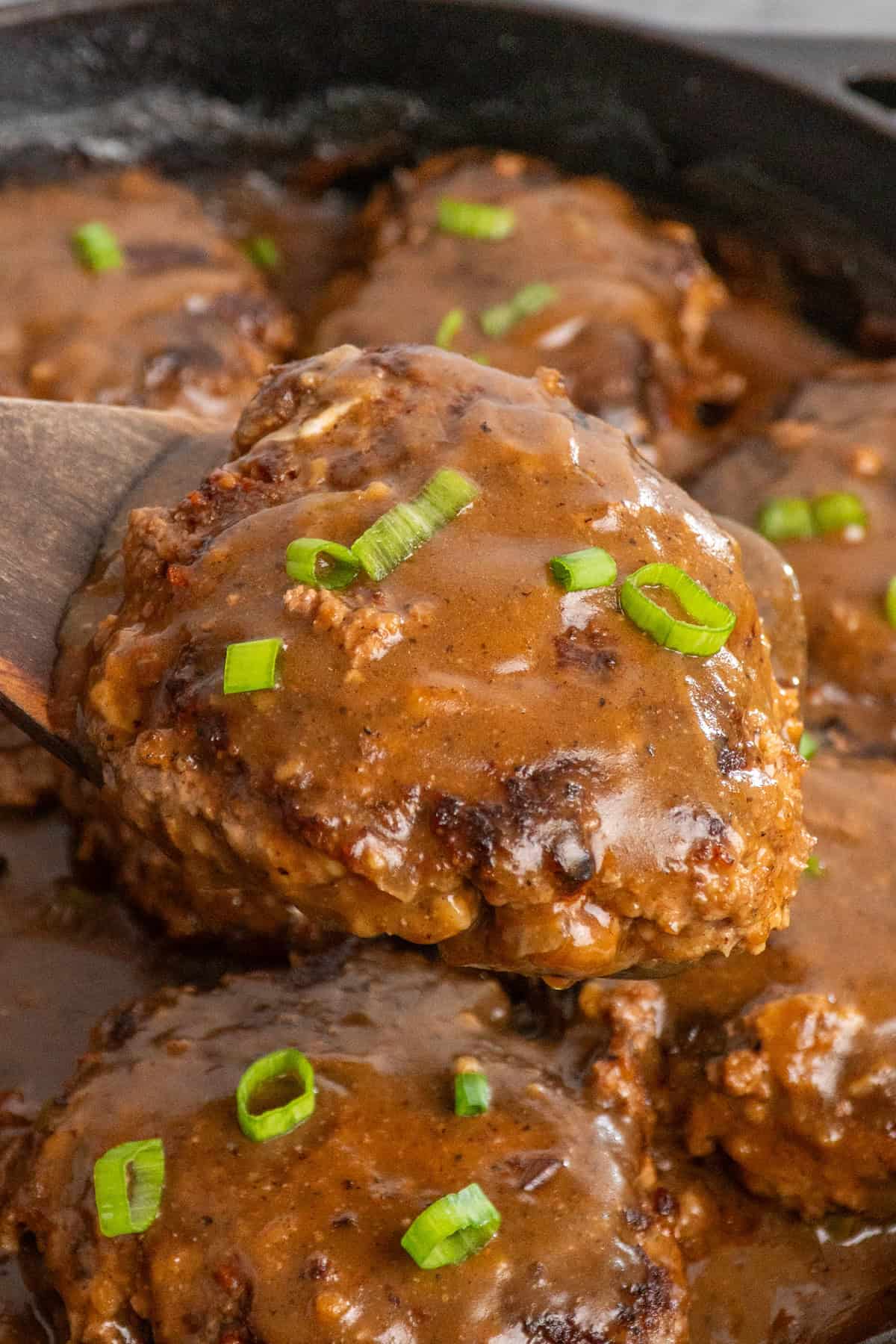 A wooden spatula holding a Salisbury steak over a skillet.