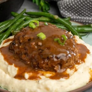 Salisbury steak with gravy on a plate of mashed potatoes with green beans.