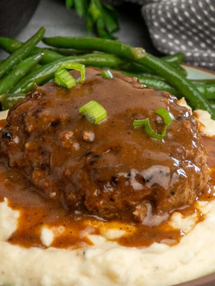 Salisbury steak with gravy on a plate of mashed potatoes with green beans.