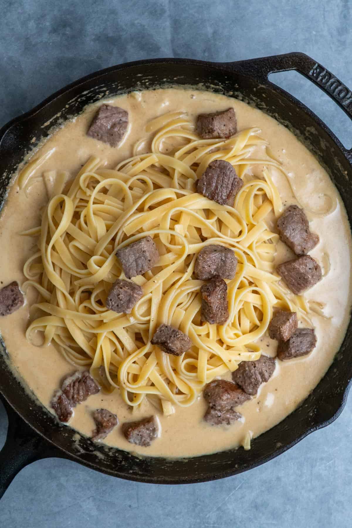 Cooked fettuccine and steak bites in alfredo sauce in a cast iron skillet.