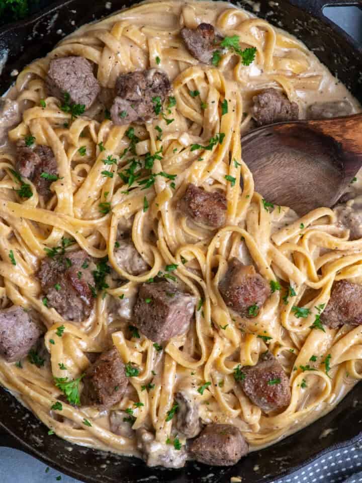 Fettuccine Alfredo with steak in a skillet and a wooden spoon to serve it.