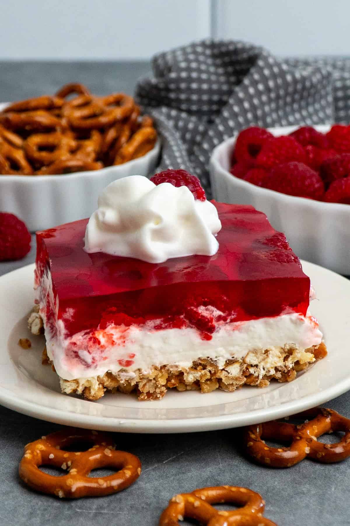 Raspberry pretzel salad on a plate with pretzels and raspberries in bowls behind it.