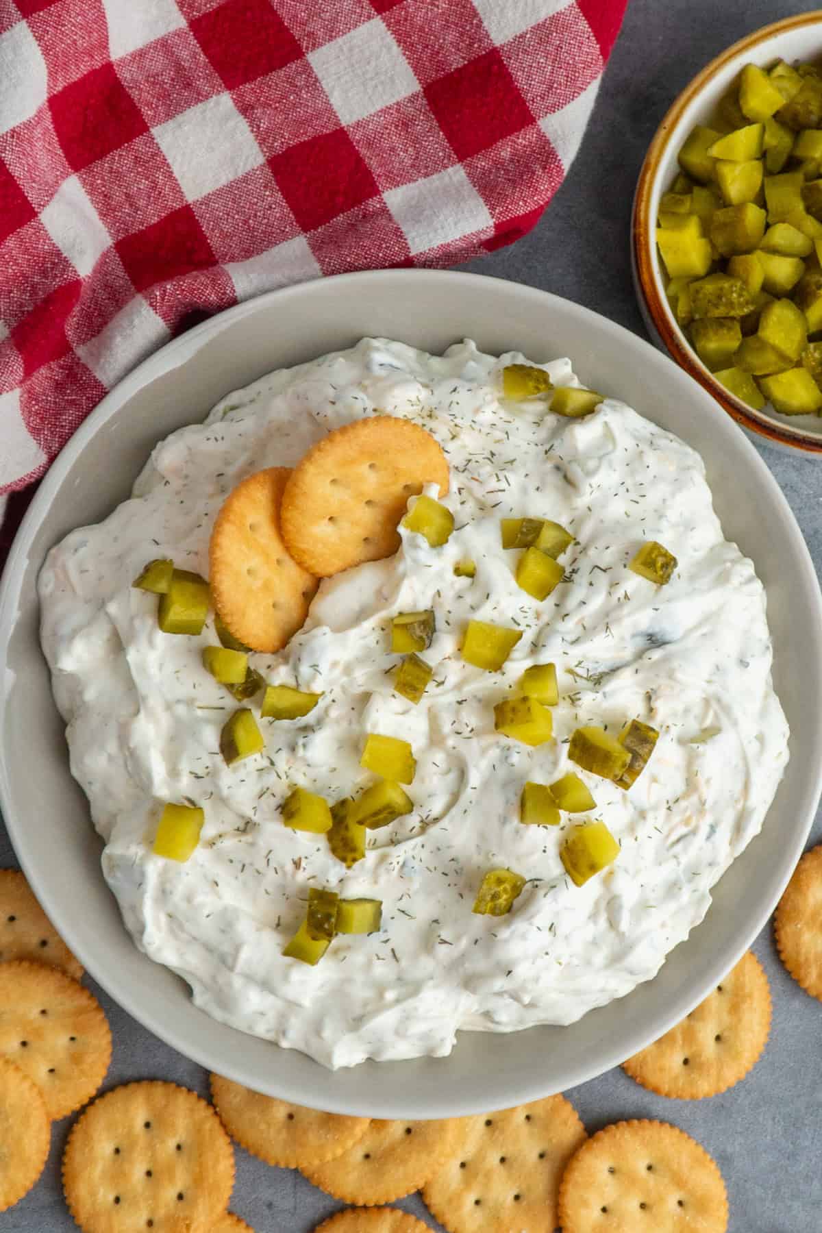 A bowl of dill pickle dip with pickles on top and crackers.