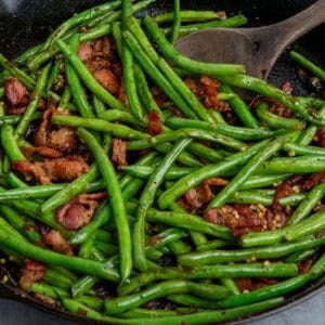 Cooked green beans with bacon in a skillet with a wooden spoon.
