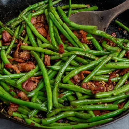 Cooked green beans with bacon in a skillet with a wooden spoon.