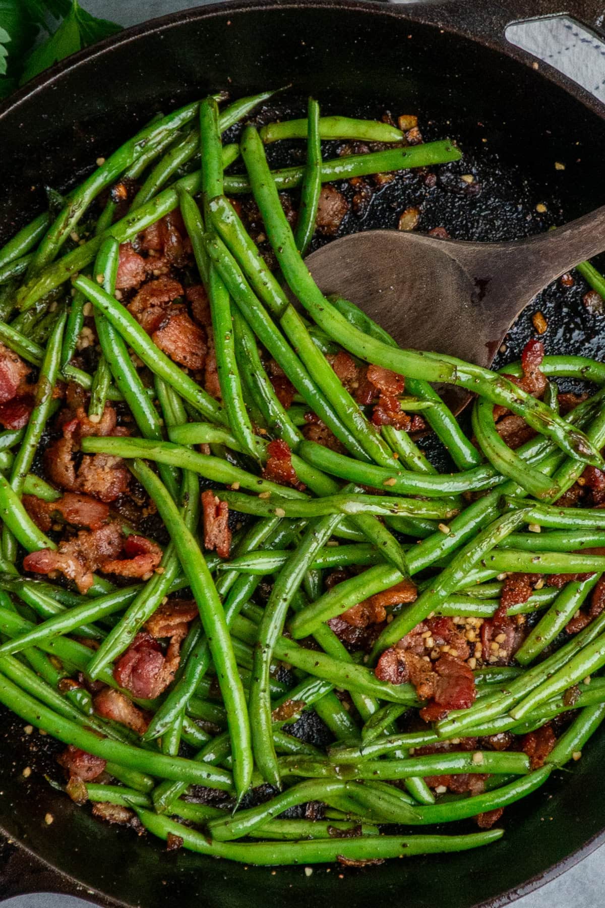 Cooked green beans with bacon in a skillet with a wooden spoon.