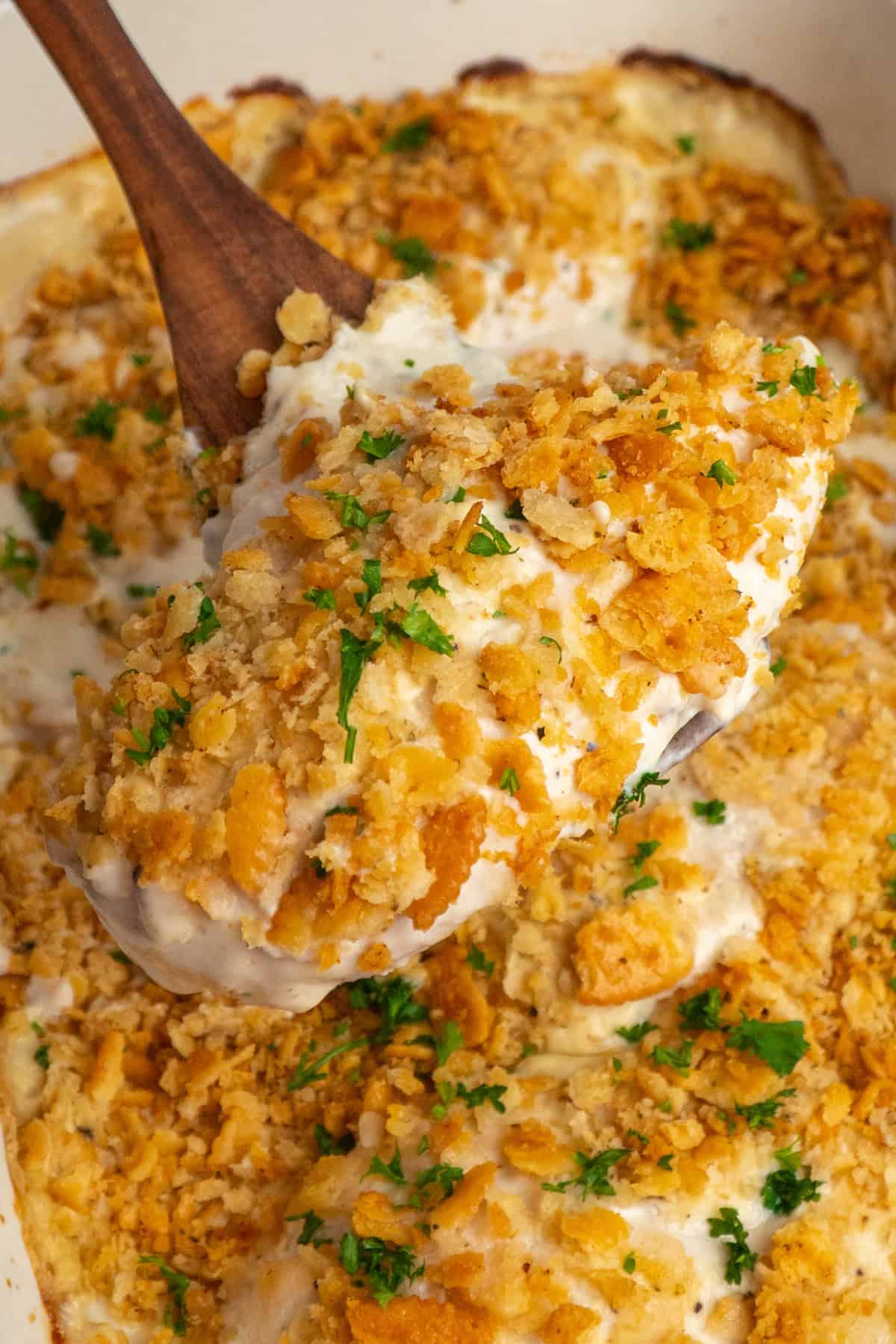 A wooden spoon scooping out some chicken with ritz cracker topping from the dish below.