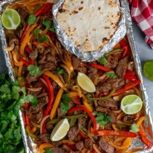 Sheet pan steak fajitas with limes all around and tortillas wrapped in foil.
