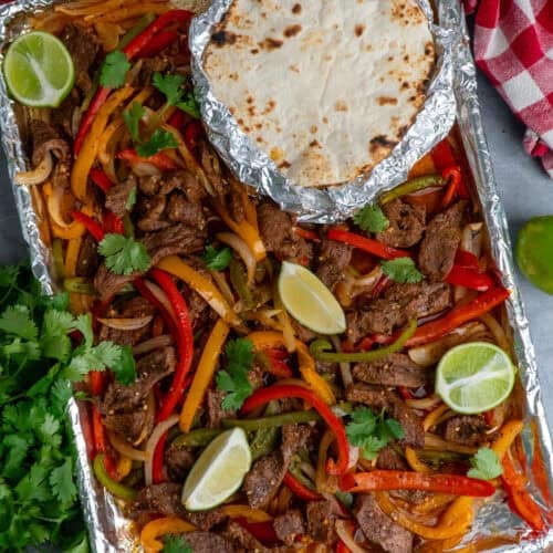 Sheet pan steak fajitas with limes all around and tortillas wrapped in foil.