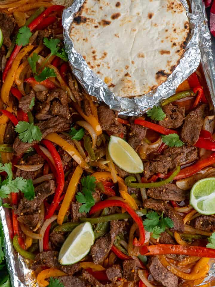 Sheet pan steak fajitas with limes all around and tortillas wrapped in foil.