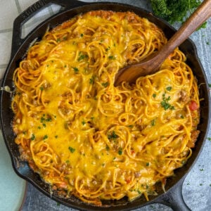Cheesy baked spaghetti in a skillet with a wooden spoon in it.
