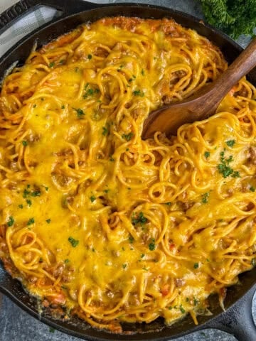 Cheesy baked spaghetti in a skillet with a wooden spoon in it.
