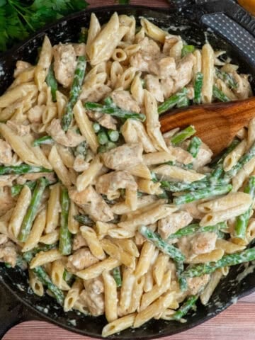 Chicken asparagus pasta in a cast iron skillet with a wooden spoon in it.