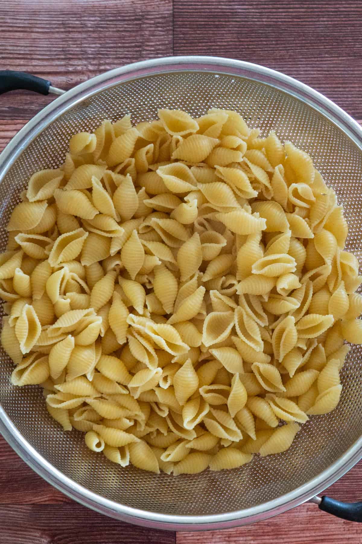 Cooked shell pasta in a strainer on a wooden countertop.