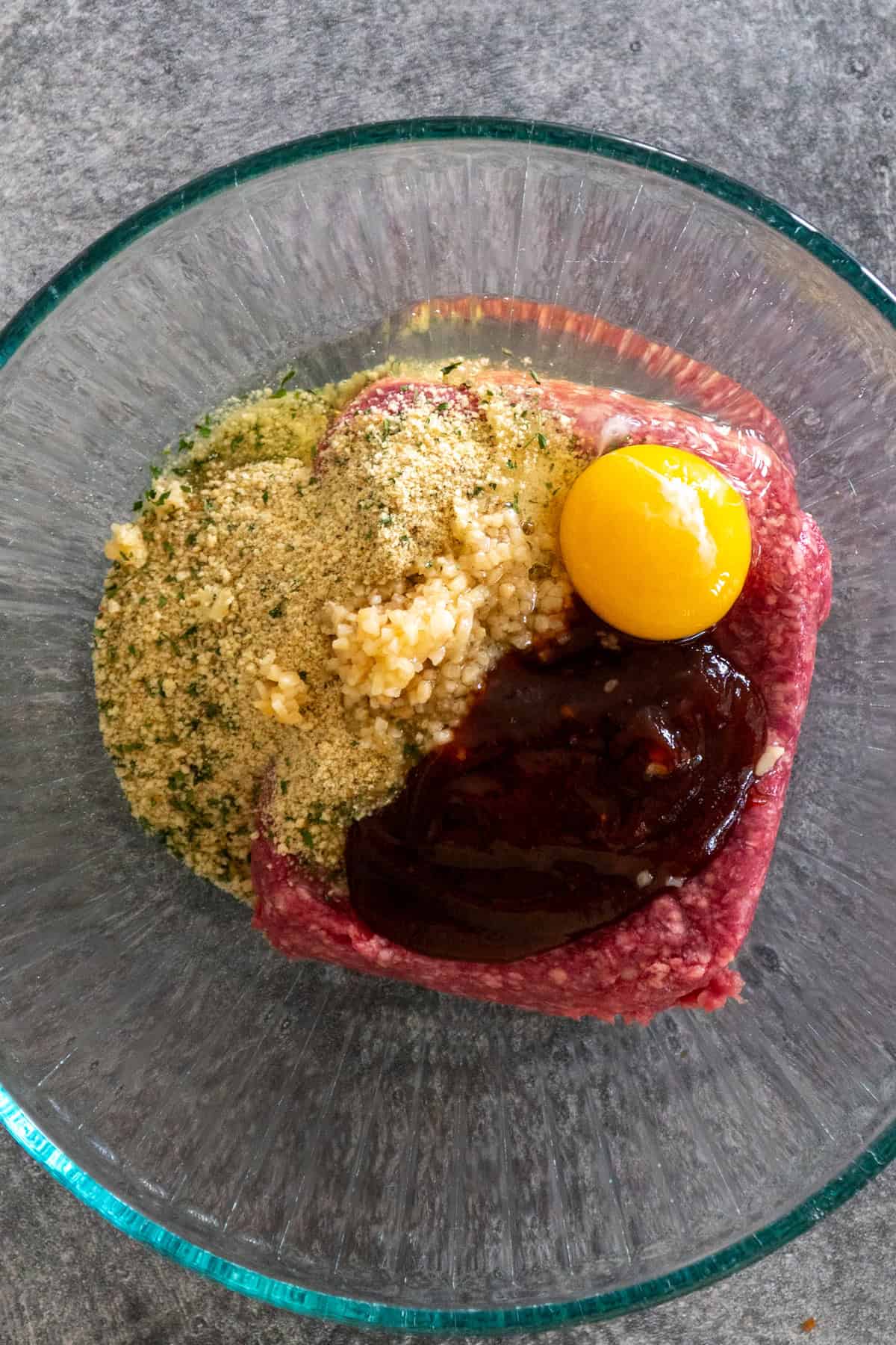 Ground beef, bbq sauce, bread crumbs, garlic and an egg in a glass bowl.