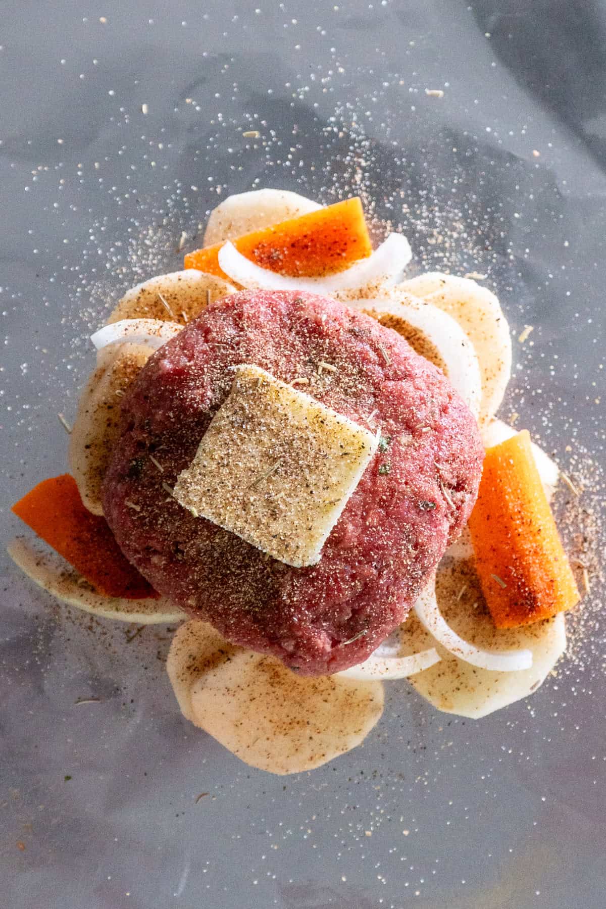 Beef patty, butter and seasonings placed on top of potatoes and veggies.